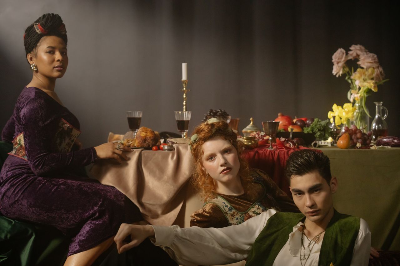 Three elegantly dressed individuals in a Renaissance-inspired setting with food and wine on a table.