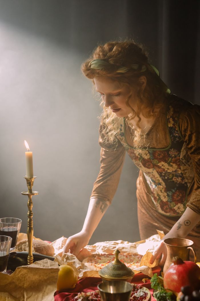 Services A woman in baroque attire sets a candlelit table with rustic food, creating a moody dining atmosphere.